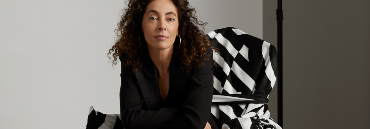 Portrait of Zahava Elenberg in a crisp black suit seated on a boldly patterned armchair, set in photography studio