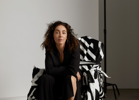 Portrait of Zahava Elenberg in a crisp black suit seated on a boldly patterned armchair, set in photography studio